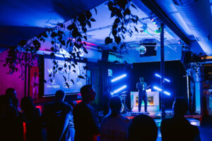 Emcee speaking on stage at a live event with vibrant lighting and audience in foreground. POC