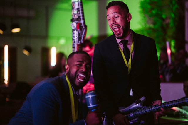 Two Colorado event pros from a local DJ company laugh and perform with a toy mic and inflatable guitar at a vendor networking party—showcasing the energy, personality, and partnership behind great vendor-venue collaboration.