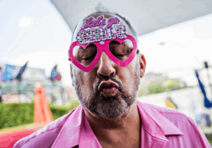 Carlo Stuart laughing and being playful at the Pink Pony Club party, wearing a bright pink shirt and oversized novelty glasses.